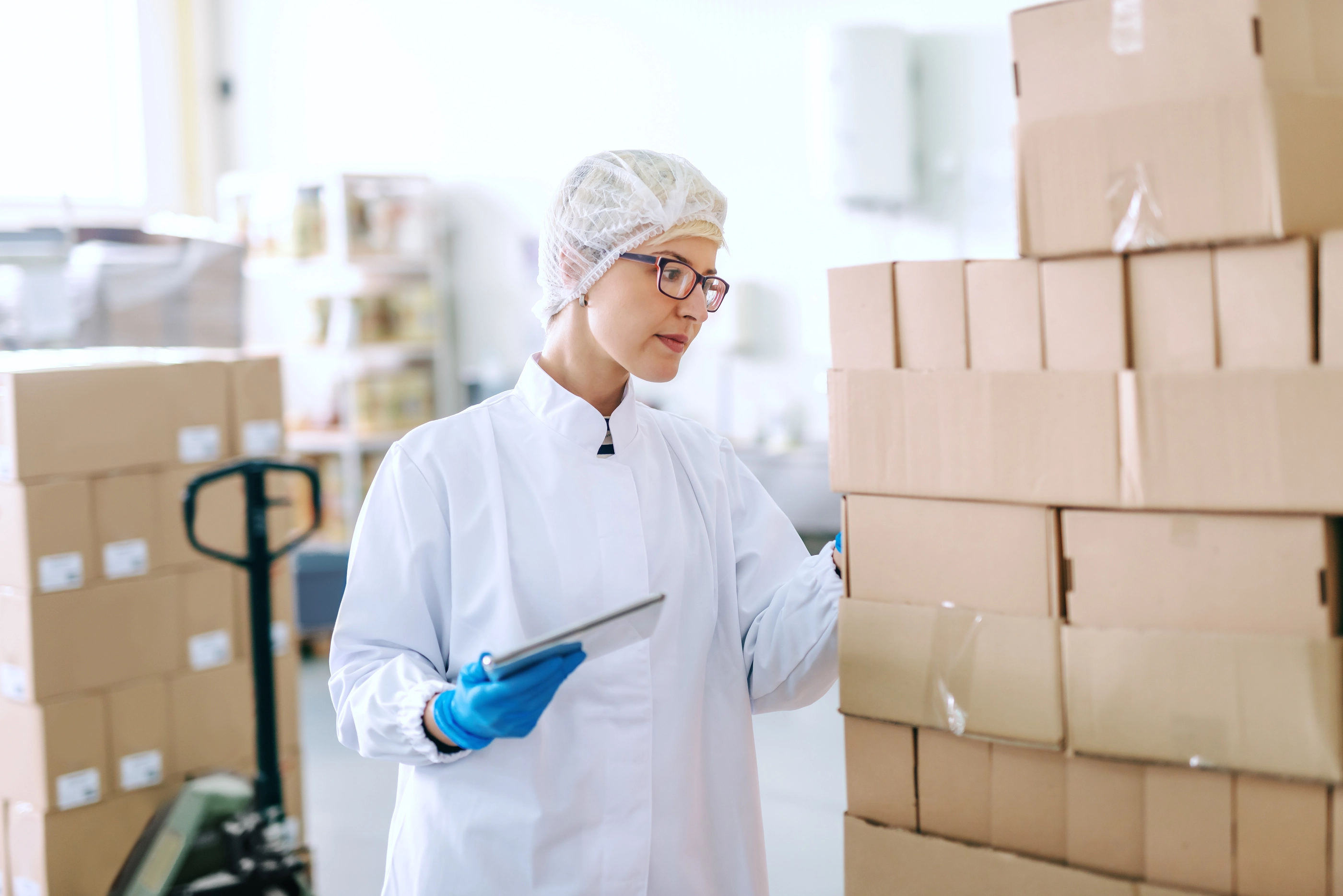 Caucasian blonde employee in sterile uniform standing and dealing with logistic of products. Food factory interior. In hand tablet.